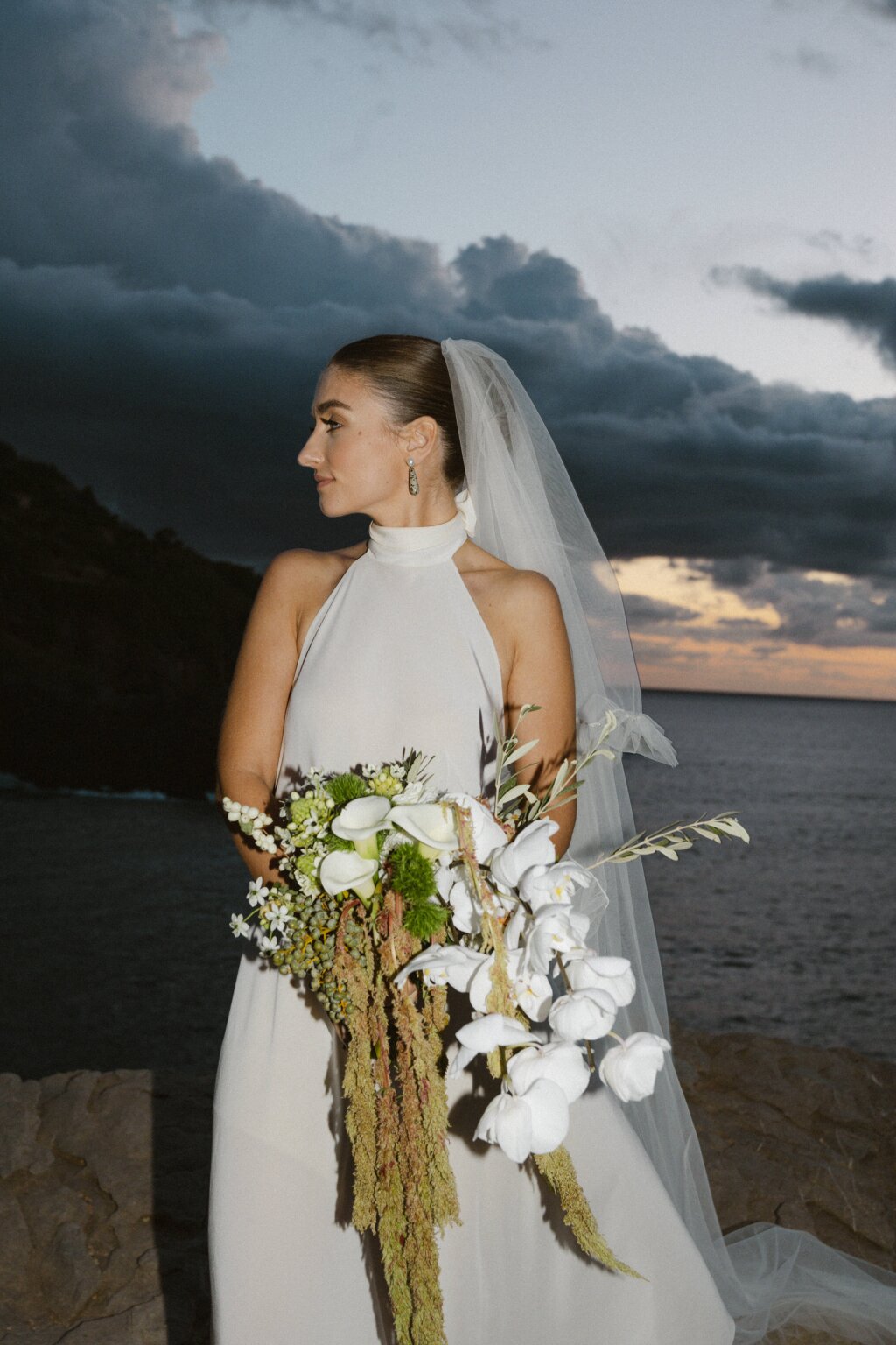 Die Braut steht mit einem prachtvollen Orchideenstrauß am Meer, umgeben von dramatischem Himmel beim Sonnenuntergang.