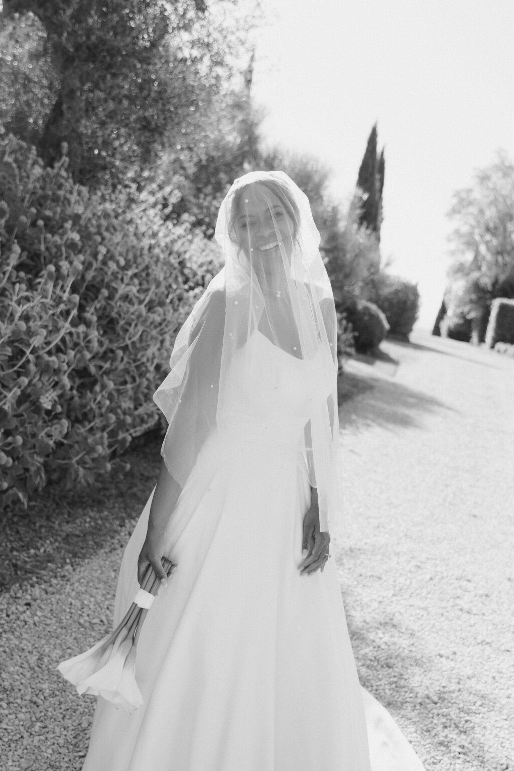 Lächelnde Braut mit Schleier und Calla-Lilien in der Hand, eingefangen in einem zarten Schwarz-Weiß-Foto unter mediterraner Sonne.