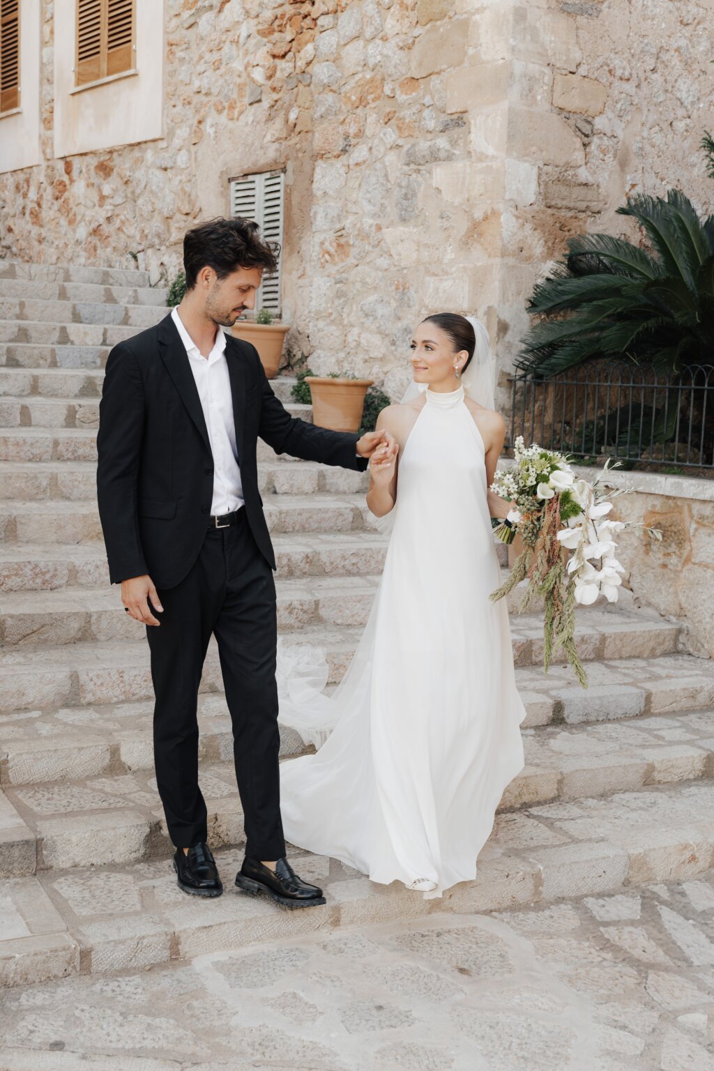 Das Brautpaar schreitet Hand in Hand eine mediterrane Steintreppe hinunter – ein eleganter Moment unter freiem Himmel.