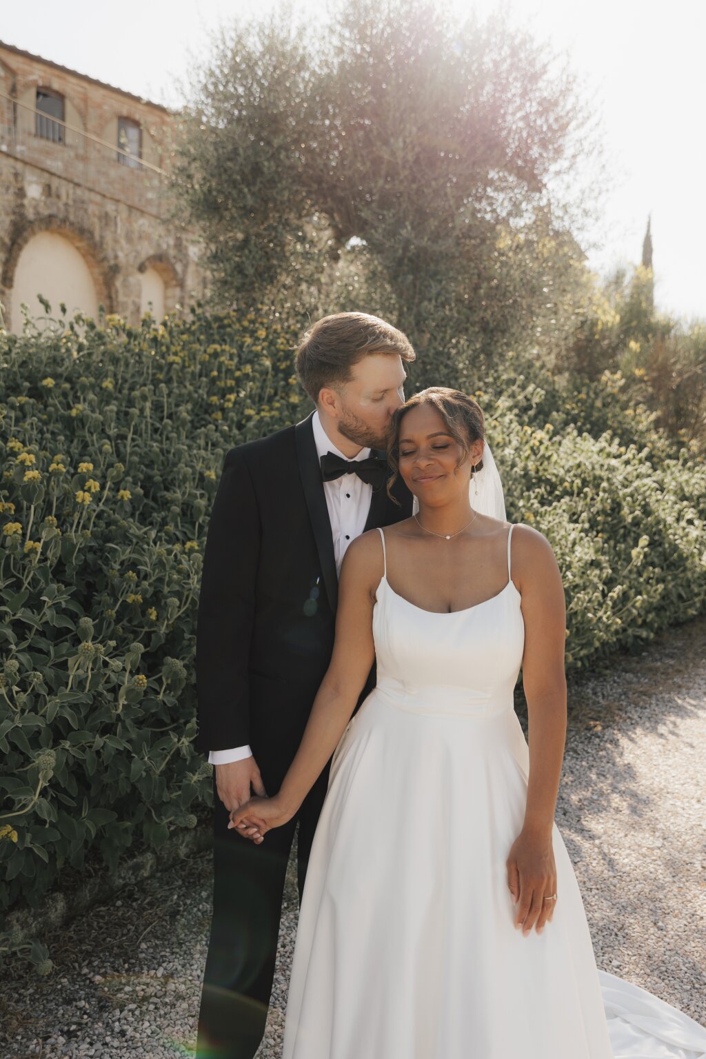 Der Bräutigam küsst zärtlich die Stirn der Braut, während sie sich vor mediterraner Kulisse liebevoll an den Händen halten – ein romantischer Moment bei ihrer Hochzeit.