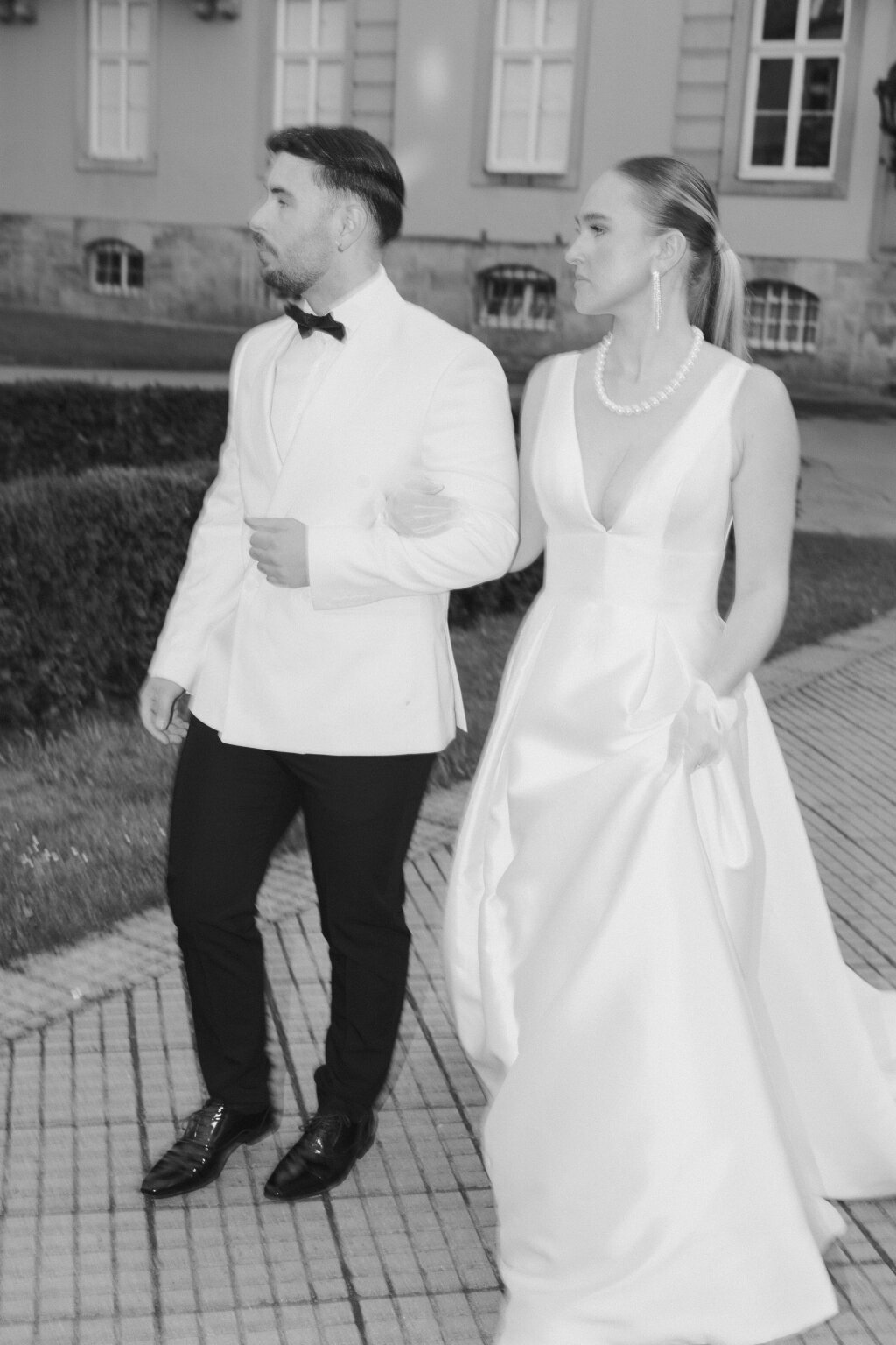 Elegantes Brautpaar schreitet stilvoll Arm in Arm entlang einer historischen Location – ein klassischer Schwarz-Weiß-Moment voller Anmut.