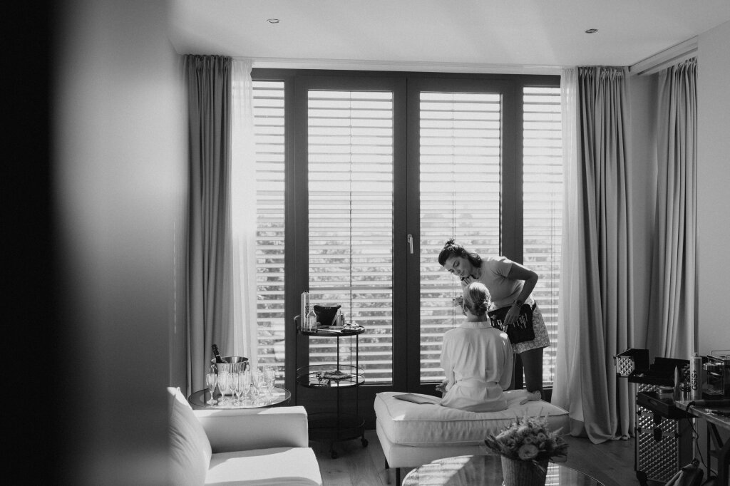 Visagistin schminkt Braut im hellen Hotelzimmer vor Panoramafenster.