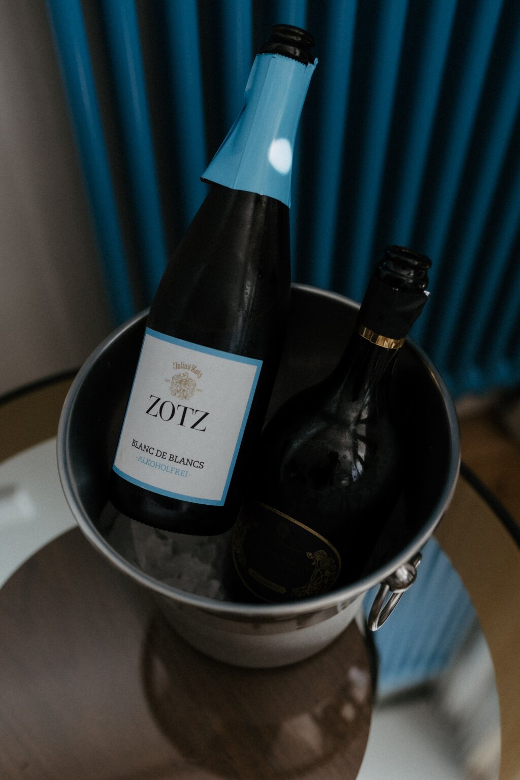 Zwei Flaschen alkoholfreier Sekt in einem Kühler auf einem Glastisch.