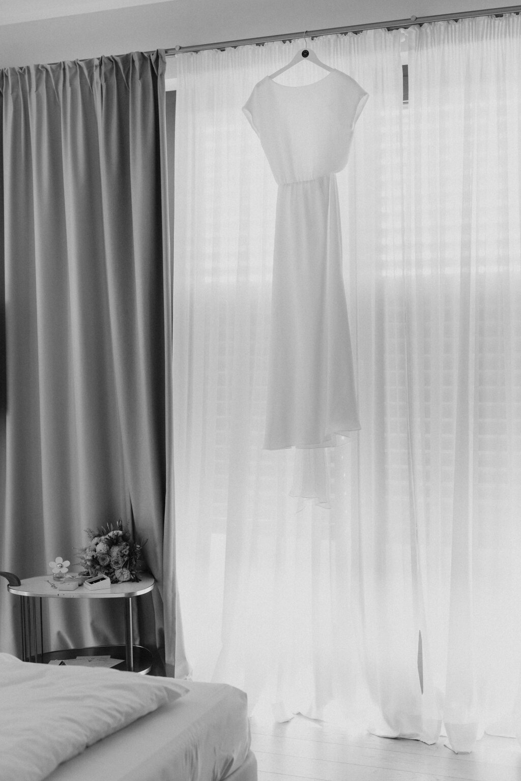 Brautkleid hängt im Hotelzimmer vor lichtdurchflutetem Vorhang.